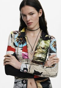 Šarena košulja dugih rukava s apstraktnim uzorcima, koja ima zatvarač na ovratniku. Uključuje crnu choker ogrlicu i srebrni privjesak u obliku srca.