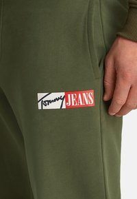 Pantalones joggers verdes oliva con un acabado texturizado, que cuentan con una etiqueta rectangular de logotipo en blanco y rojo, mostrando "Tommy JEANS" en letras gruesas.