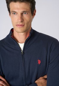 Maglione zip-up navy realizzato in tessuto a maglia con colletto a coste. Presenta un logo ricamato rosso sul petto e una fodera interna rossa.