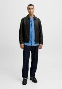 Black leather jacket over a light blue denim shirt, paired with dark blue jeans and black shoes, standing against a plain background.