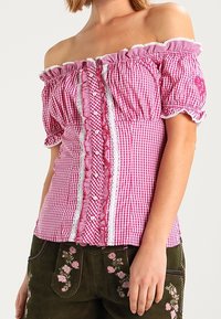 Blusa off-shoulder a righe rosa e bianche con bordi in pizzo, bottoni, maniche a sbuffo, indossata con pantaloni ricamati verdi scuri.
