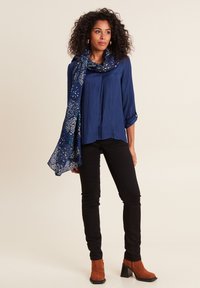 Blusa blu navy con maniche a tre quarti, abbinata a jeans skinny neri e stivali a tronchetto con tacco marroni. La sciarpa presenta un motivo floreale blu.