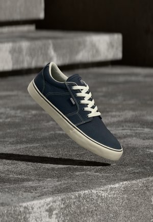Marineblauer Sneaker mit weißen Schnürsenkeln und Sohle, der über strukturierten Betontreppen im natürlichen Licht schwebt.