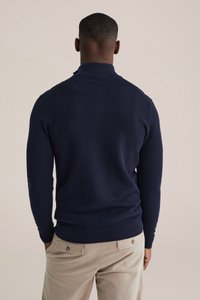 Tumši zils adīts džemperis ar augstu apkakli un ribas aprocēm, ar teksturētu rakstu. Valkāts kopā ar gaiši bēšām biksēm.