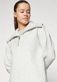 Ανοιχτό γκρι πουλόβερ quarter-zip από υφασμάτινη υφή με μικρές μαύρες κουκίδες. Διαθέτει ψηλό γιακά και κρεμαστούς ώμους.