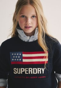 Maglione blu navy con motivo bandiera degli Stati Uniti rossa e blu, logo "SUPERDRY" bianco e rosso, indossato sopra una camicia a righe azzurre e bianche con colletto.