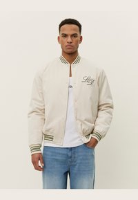 Jachetă bomber bej cu manșete și guler ribbed în dungi verzi. Prezintă un logo pe piept, material neted și închidere frontală cu capse.
