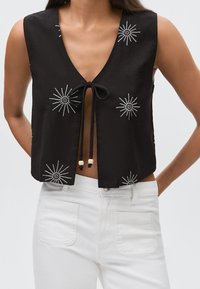 Top corto senza maniche nero con ricamo a stella bianca, annodato davanti con due cordini neri pendenti con perline in legno, abbinato a pantaloni bianchi.