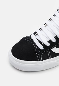 Zwarte sneaker met een suède neuskap, witte veters en rubberen zool. Het ontwerp bevat witte stiksels en een logo-accent aan de zijkant.