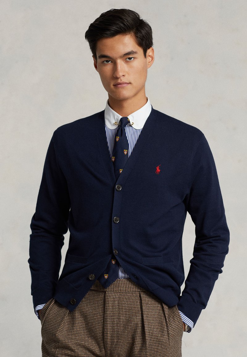 Cardigan blu navy realizzato in tessuto lavorato a maglia, con scollo a V profondo, bottoni frontali e due tasche, indossato sopra una camicia a righe e una cravatta.