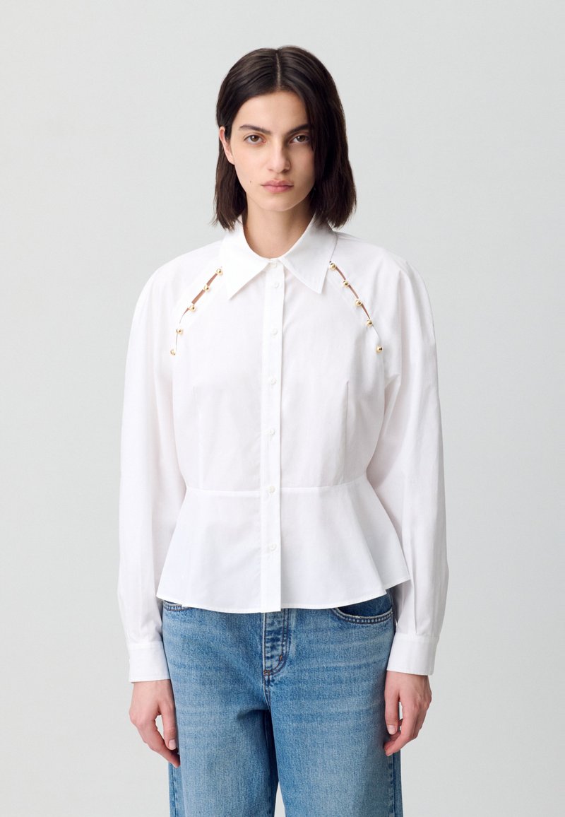 Modella che indossa una camicia bianca con dettagli dorati sulle spalle, abbinata a jeans blu, su uno sfondo grigio chiaro.