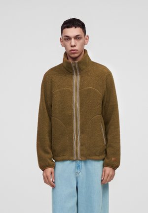 Un homme porte une veste en polaire marron avec un col montant et une fermeture éclair devant, associée à un jean bleu clair sur un fond gris uni.