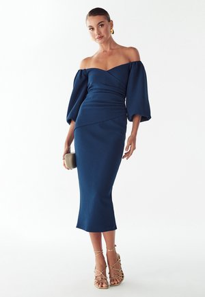 Vestido azul marino con escote barco, mangas abullonadas, cuerpo ajustado y largo midi. Lleva sandalias de tiras y un pequeño bolso de mano beige.