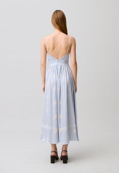 Donna con capelli lunghi e lisci, indossa un abito midi floreale azzurro chiaro con spalline sottili e sandali neri con tacco alto, in piedi di spalle.
