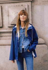 Ženska z dolgimi svetlo rjavimi lasmi, oblečena v večplastno modro jeans jakno in svetlo modro nepremočljivo jakno, stoji ob teksturirani kamniti steni.