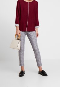 Femme portant un haut à manches longues bordeaux, un pantalon slim à carreaux, des mocassins noirs, tenant un sac beige avec des poignées en bois devant un fond blanc.