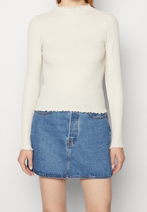 Femme portant un haut crème ajusté à manches longues avec des finitions effet feuille de laitue et une mini-jupe en jean bleu avec une couture devant et des poches.