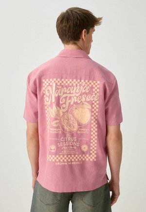 Mężczyzna ubrany w różową koszulkę z krótkim rękawem z grafiką cytrusów w stylu vintage i napisem na plecach, w połączeniu z szarymi spodniami.