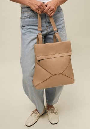 Persona con vaqueros azul claro y zapatos tipo mocasín beige sosteniendo un gran bolso de mano de cuero marrón claro con patrones de costuras geométricas.