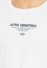 Bílé tričko s černým textem "ALPHA INDUSTRIES" a menším textem o dodavateli ministerstva obrany USA, spolu s logem hvězdy níže.