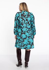 Blumenkleid mit einem türkisfarbenen und schwarzen Muster, langen Ärmeln und einem Schlüsselaugen-Rücken. Kombiniert mit glänzenden, knielangen braunen Stiefeln.
