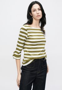 Olīvzaļš un balts svītrains krekls ar garām piedurknēm un laivas izgriezumu, piedurknes nolocītas uz augšu, kombinēts ar tumšas krāsas džinsa biksēm.