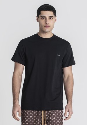 Junger Mann mit kurzen dunklen Haaren, der ein schlichtes schwarzes T-Shirt und gemusterte braune Shorts trägt, steht vor einem einfarbigen hellen Hintergrund.