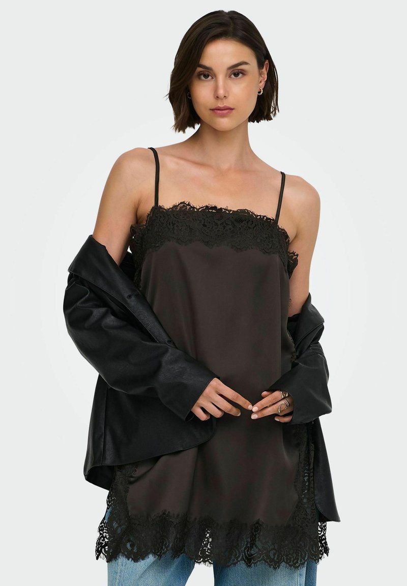 Jeune femme portant un top noir à bordure en dentelle, une veste noire ample tombant des épaules, un jean bleu et des bagues en or aux doigts.