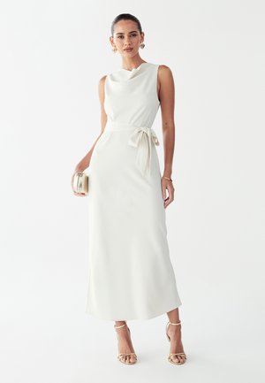 Vestido largo blanco sin mangas con corpiño ajustado, detalle de lazo en la cintura y una falda lisa y fluida. Modelo sosteniendo un clutch metálico.