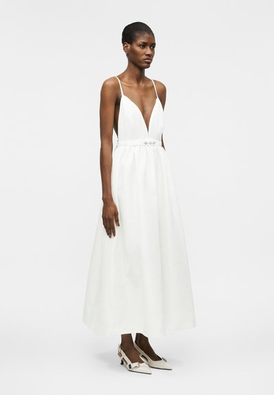 Γυναίκα που φοράει λευκό μίντι φόρεμα χωρίς μανίκια με βαθύ V-ντεκολτέ και διακοσμητική μέση, σετ με λευκά μυτερά παπούτσια με χαμηλό τακούνι.