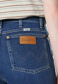 Mörkblå denimjeans med en läderpatch från Wrangler på bakfickan, prydda med zickzackstygn och metallnitar.