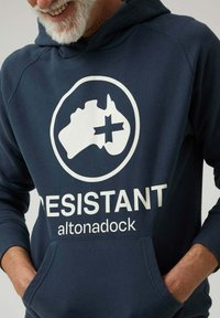 Sudadera con capucha azul marino hecha de tela suave, con un gran gráfico blanco de Australia con una cruz, y el texto "RESISTANT altonadock" debajo.