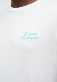 Κοντινό πλάνο ενός ατόμου που φοράει ένα λευκό πουκάμισο με κεντημένο το κείμενο "Tommy Hilfiger" σε τιρκουάζ στο στήθος.