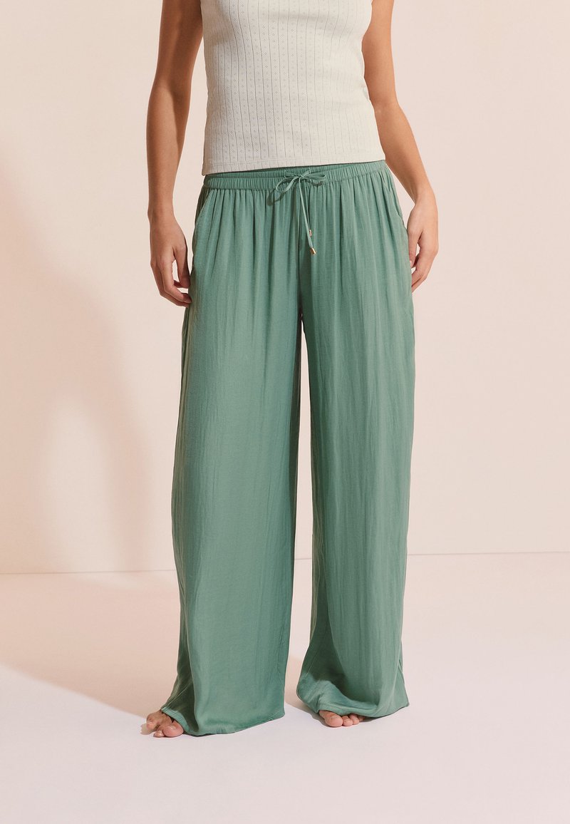 Mujer vistiendo pantalones anchos de pierna amplia y color verde claro con cordón en la cintura y una camiseta sin mangas blanca acanalada, de pie descalza sobre un suelo liso.