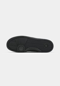 Svart yttersåle til sneakers med en teksturert gummioverflate, som har zigzag-mønstre og sirkulære dekkelementer. Merkenavnet er graverte langs kanten.