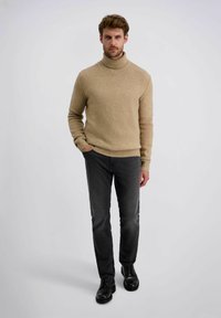 Mann steht mit den Händen in den Taschen, trägt einen beigen Rollkragenpullover, schwarze Jeans und schwarze Schnürschuhe vor einem einfarbigen Hintergrund.