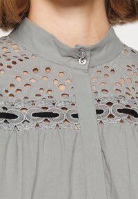 Camisa gris claro con un alto cuello, que presenta intrincados diseños recortados y detalles bordados. Cierre de botones con acentos de cinta negra.