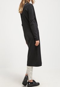 Donna che indossa un lungo vestito nero, leggings texturizzati chiari e scarpe platform nere lucide, in piedi davanti a uno sfondo semplice.