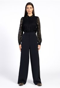 Vrouw staat in een zwarte doorschijnende blouse met lange mouwen en pailletten, hoge taille wijd uitgesneden zwarte broeken en zwarte schoenen met studs.