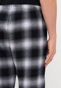 Pantalons à carreaux noirs et gris avec une texture douce, présentant un léger brillant et une taille à cordon de serrage. Le design inclut des motifs de carreaux contrastants.