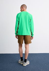 Långärmad grön skjorta med bruna detaljer, kombinerad med bruna shorts. Våffelstruktur på skjortan, avslappnad passform. Modellen står med ryggen mot kameran.