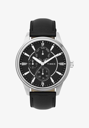 Timex sat s crnim teksturiranim brojčanikom, kućištem od srebrnog nehrđajućeg čelika, bijelim oznakama sati i crnim kožnim remenom. Ima prikaz datuma i dana.