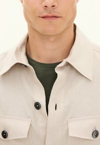 Lys beige button-up skjorte med tekstureret stof, to forlommer og mørke knapper over en grøn crew neck t-shirt.