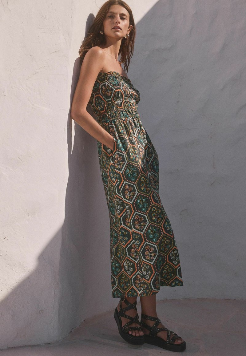 Groene jumpsuit met een honingraatpatroon, elastische halslijn, zakken, en gecombineerd met zwarte sandaaltjes met banden. Glad textuur van de stof.