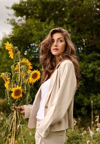 Jeune femme aux longs cheveux ondulés tenant un bouquet de tournesols, portant une veste de costume beige et un haut blanc en extérieur avec un fond de feuillage vert.