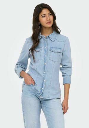 Woman with long dark hair wearing light blue denim shirt and jeans, standing with one hand in pocket, looking slightly to the side.