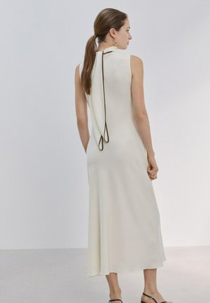 Femme avec une queue de cheval brune portant une robe midi blanche sans manches avec un détail d'écharpe bordée de noir à l'arrière, debout devant un fond clair uni.