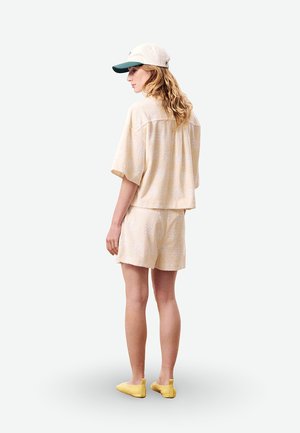 Femme debout de dos, portant une chemise à manches courtes texturée beige et un short, une casquette blanche et verte, et des chaussures jaunes sans lacets.