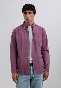 Langärmeliges Hemd in gedämpftem Mauve mit Brusttasche, Kragen und geradem Saum. Wird über einem weißen T-Shirt getragen und mit hellen Jeans kombiniert.