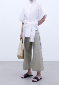 Witte, kortom mouwen shirt met een striktaille, gecombineerd met gestructureerde, wijde beige broek en zwarte instappers. Licht handtasje in de hand.